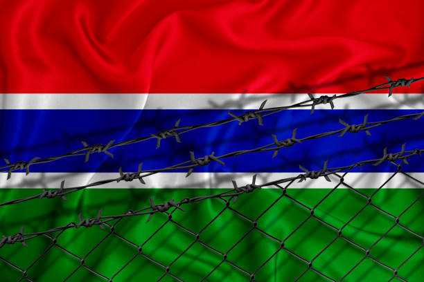 istock Gambia flag development, fence mesh and barbed wire. Emigrants isolation concept. With place for your text. 1289727598