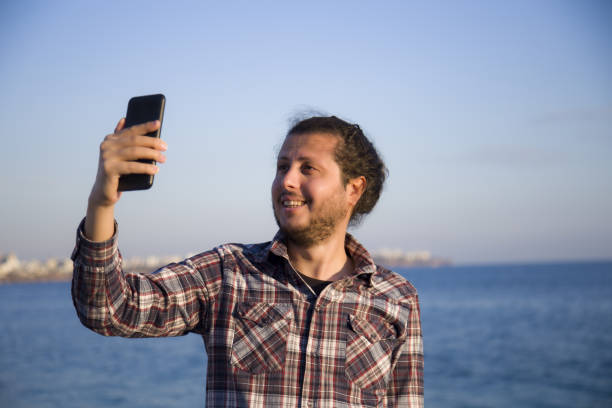 istock Young man on vacation laughing at the beach taking selfie 1289727897