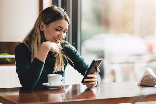 Young happy woman at restaurant drinking coffee and using mobile phone.