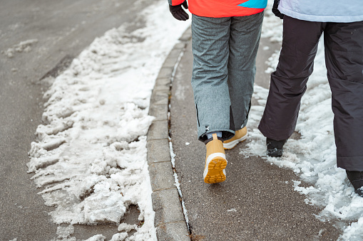 Legs of two children walking in warm clothes on the sidewalk in a snowy winter time.
