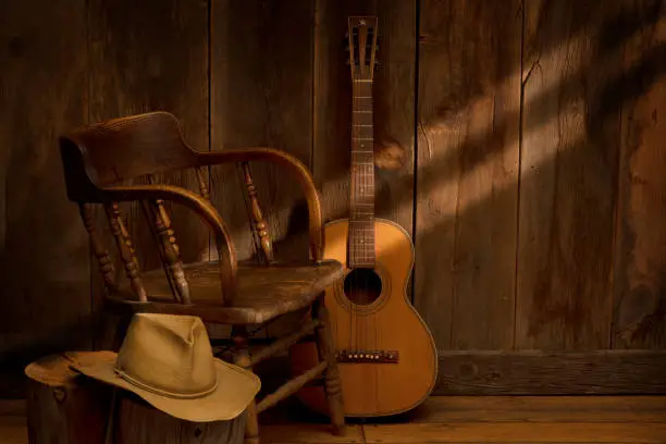 The image is a Wild West still life with barn wood background. A vintage saloon chair is placed next to a vintage guitar on one side and cowboy hat on the other. The scene has subdued lighting with sun streaks streaming in from the side.