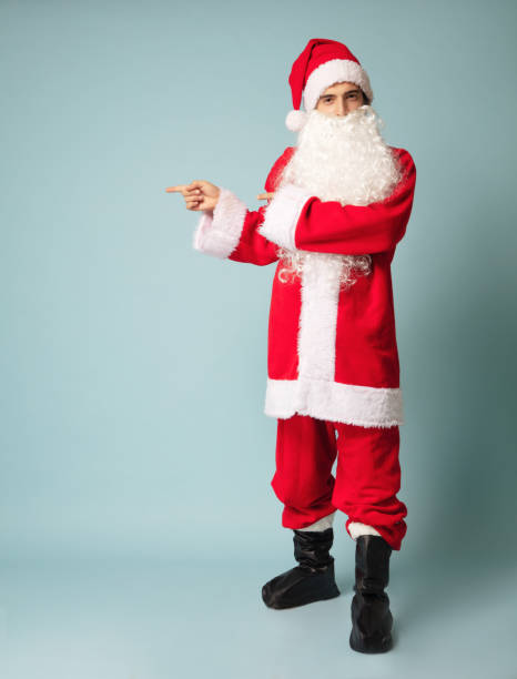 istock Young Santa Claus pointing side with fingers front of blue background. 1289747112