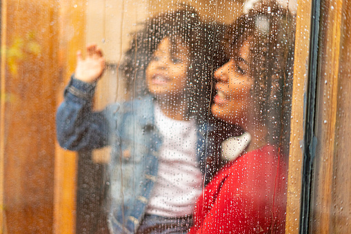 mom Latin woman of average age of 29 years old with brown skin and afro hair carries her little daughter of 5 years old with afro hair and brown skin both are looking out the window of their house while outside it rains and the drops fall on the glass