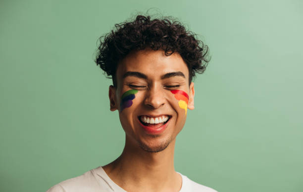 Cheerful gay man with rainbow face paint Close-up of a cheerful young man with pride flag painted on face. Gay man with rainbow face paint smiling against pastel green background. gender fluid photos stock pictures, royalty-free photos & images