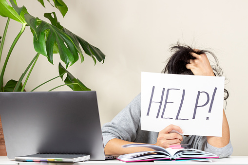 Tired frustrated woman sitting at table and holding card with word Help. Online working, learning from home.