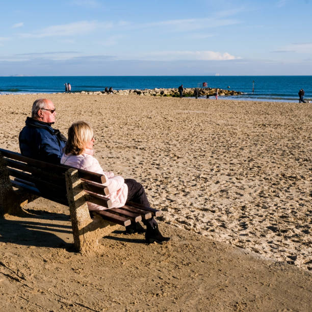 Senior Older Man and Woman Couple Sitting On A Wooden Bench Relaxing Looking Out To Sea Poole Dorset UK, December 06 2020,  Senior Older Man and Woman Couple Sitting On A Wooden Bench Relaxing Looking Out To Sea sandbanks poole harbour stock pictures, royalty-free photos & images