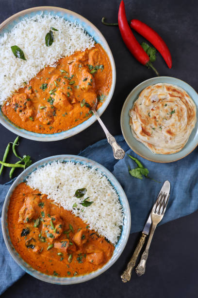 Stock photo showing elevated view of blue plates of Butter chicken breast chunks sauce and white rice, served with lachha paratha (layered flatbread). imagen de platos azules rellenos de pollo de mantequilla / curry tikka, grandes trozos de carne de pechuga de pollo en salsa de curry con guarnición de rodajas de cebolla roja y semillas de mostaza con arroz blanco, lachha paratha (pan plano en capas), fo