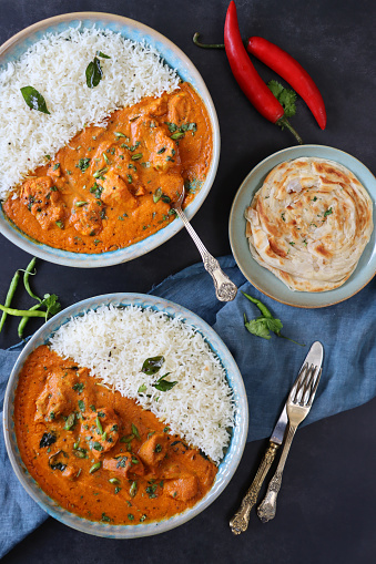 Stock photo showing elevated view of blue plates of Butter chicken breast chunks sauce and white rice, served with lachha paratha (layered flatbread).