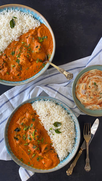 Stock photo showing elevated view of blue plates of Butter chicken breast chunks sauce and white rice, served with lachha paratha (layered flatbread). imagen de platos azules rellenos de pollo de mantequilla / curry tikka, grandes trozos de carne de pechuga de pollo en salsa de curry con guarnición de rodajas de cebolla roja y semillas de mostaza con arroz blanco, lachha paratha (pan plano en capas), fo