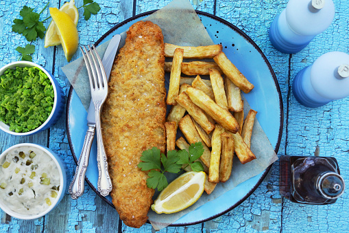 Stock photo showing an elevated view of a turquoise plate and grease proof paper that is filled with a portion of battered cod / fish and chips, with a lemon slice and garnished with parsley, besides bowls of mushy peas and tartare sauce, salt and pepper mills and a bottle of vinegar.