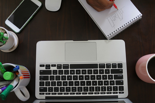 Stock photo showing an elevated view of a home office working desk with unrecognisable man drawing on a note pad. Working from home concept.