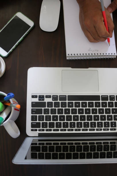 Stock photo showing an elevated view of a home office working desk with unrecognisable man writing on a note pad. Working from home concept.