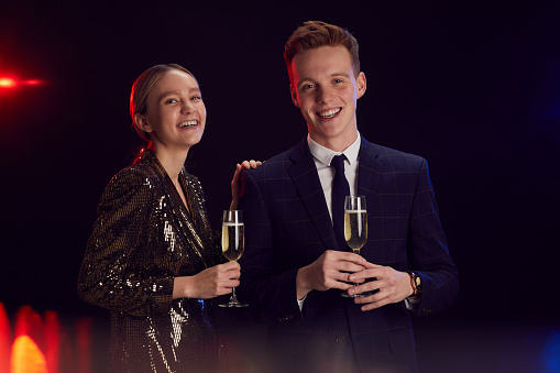 Portrait of happy young couple holding champagne glass and looking at camera while posing at party standing against black background, copy space