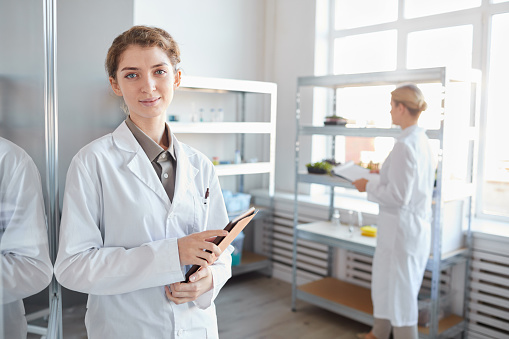 Waist up portrait of young female scientist looking at camera and holding clipboard while standing by glass wall in medical laboratory, copy space