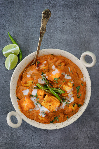 Stock photo showing an elevated view of a meal of Shahi paneer cheese curry sauce in a kadhai-style bowl on a grey surface. The dish has been garnished with fresh, green coriander leaves.