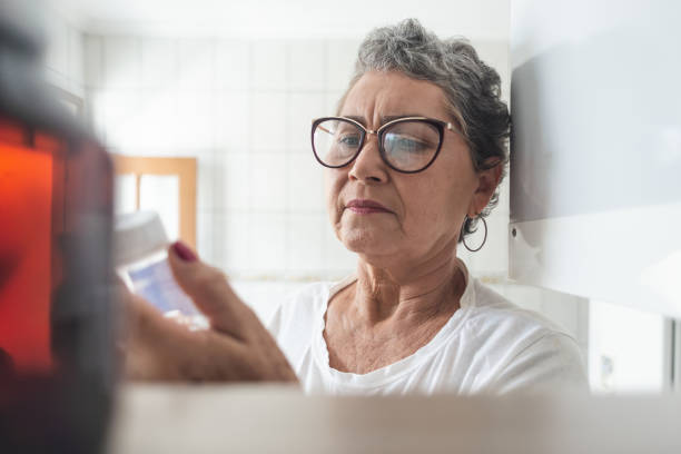 mulher idosa olhando para um frasco de medicação retirado do armário de remédios. vista do interior do gabinete - vitamin pill nutritional supplement pill bottle - fotografias e filmes do acervo