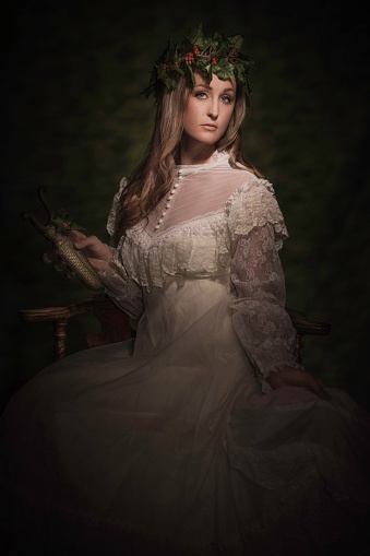 This image is a classical painterly style portrait of a young woman in white lace formalwear with a holly crown on, holding an old brass antiquity item and looking towards the camera