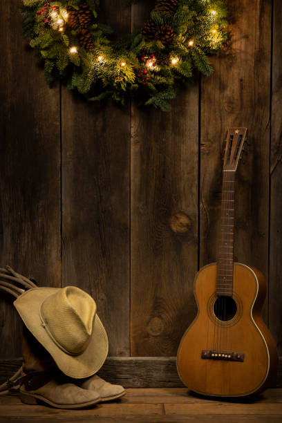 Wild West holiday theme featuring cowboy boots and hat along with a vintage guitar against a barn wood wall with partial lighted evergreen wreath. Room for overprint. No people