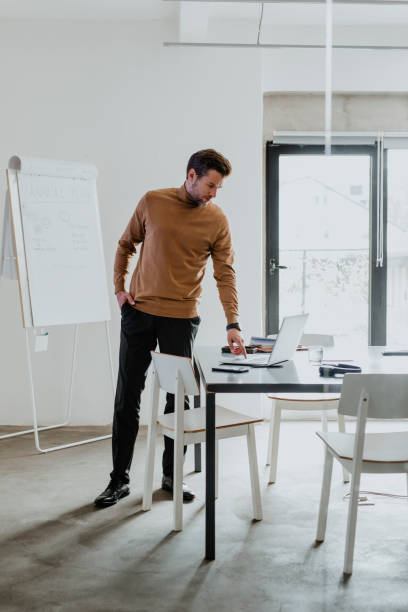 istock Young Male Teacher Standing Alone in the Classroom and Using his Laptop, Preparing for the Next Class 1289815542