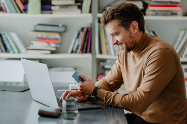 istock Smiling Young Man Wearing a Brown Turtleneck Sweater Paying his Bills on his Laptop Computer (Side View) 1289815561