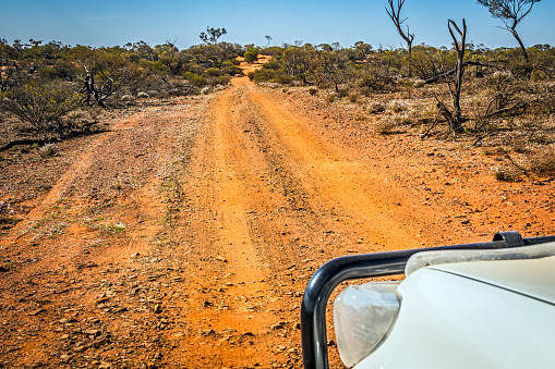 Looking over the front of a white 4X4 at a rough, red stony desert track leading through low bushes towards a sand dune in the distance: represents adversity, challenge, driving skill, resilience, in a recreational activity. Desert Road Trip.