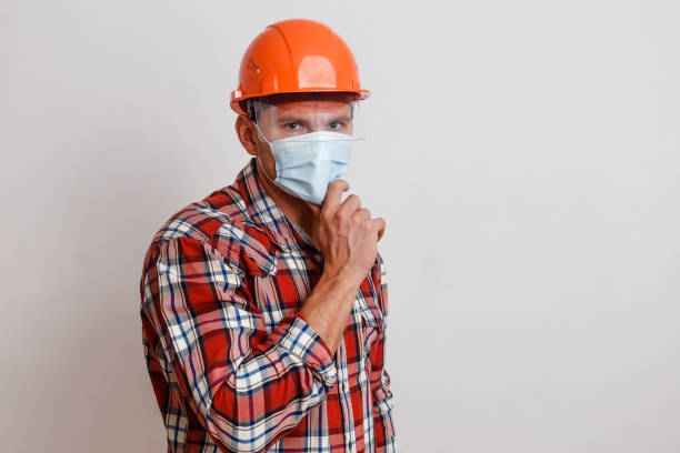 a man in a plaid shirt and an orange protective helmet on his head and wearing a medical mask on white background. mask mode. stop coronavirus a man in a plaid shirt and an orange protective helmet on his head and wearing a medical mask on a white background. mask mode. stop coronavirus tartan army stock pictures, royalty-free photos & images
