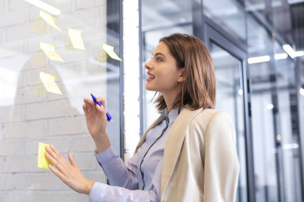 istock Attractive busness woman post stickers on glass at office and writing on them. 1289830826