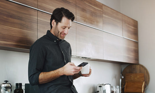 Shot of a young man using a smartphone and having coffee in the kitchen at home