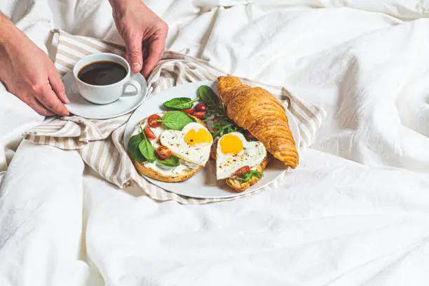 Photo of Valentine's day concept. Breakfast in bed on Valentine's Day. Sandwiches with croissant, bagel, cream cheese and fried eggs hearts on white bed background, copy space.