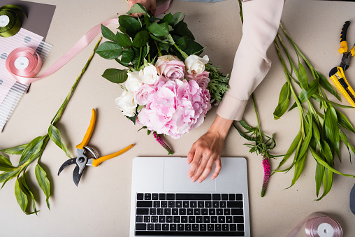 Cropped view of female florist using laptop, while holding bouquet on desk with tools, plants and decorative ribbons