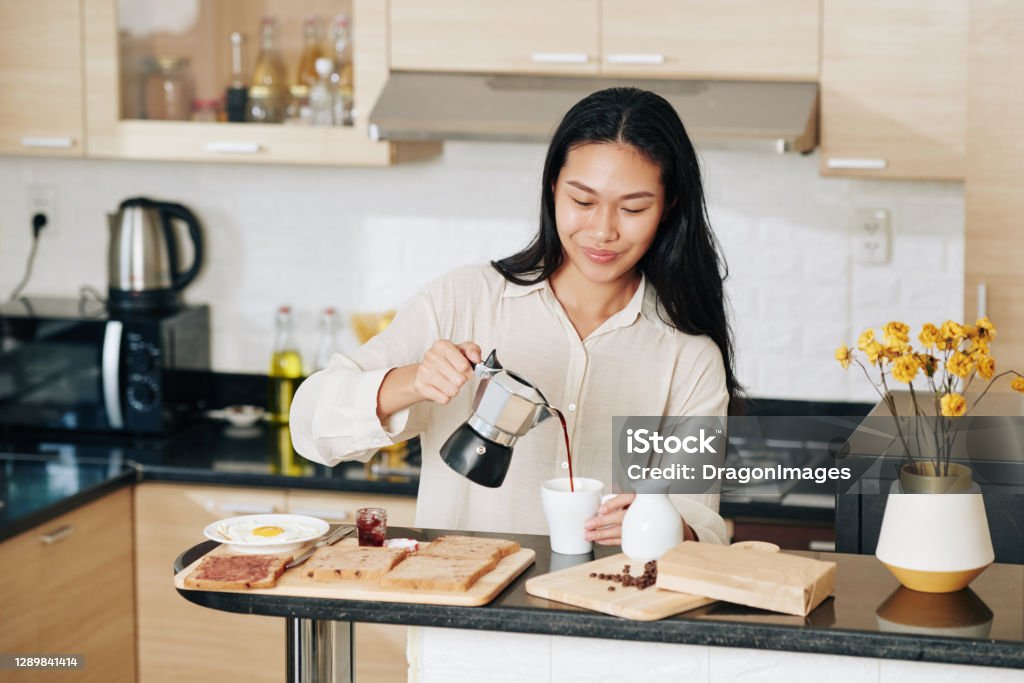 Woman pouring cup of coffee Smiling pretty Chinese woman pouring coffee in cup when eating breakfast in kitchen Coffee - Drink Stock Photo
