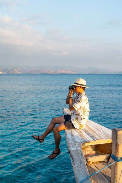 giovane viaggiatore asiatico seduto sulla punta della barca da pesca e scattare foto al bellissimo tramonto serale. un uomo che viaggia sull'isola di flores, indonesia - flores man foto e immagini stock