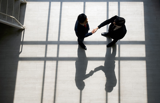 Business partners shaking hands in the modern office. Standing near the window while the sunlight shines on them. There is a shadow on the wooden floor