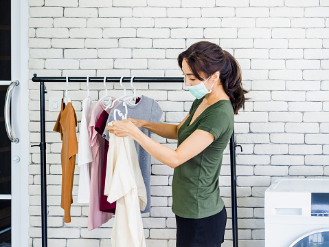 Young beautiful Asian woman, housewife in casual wearing protective face mask hanging dry shirt with hanger on clothesline after washing near wash machine in laundry room on white wall background.