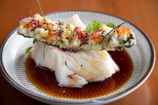Serving of sea bass with tempura stick on top in soy sauce, plate on wooden table