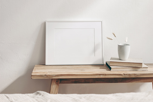 Horizontal white frame mockup on vintage wooden bench, table. Ceramic mug with dry Lagurus ovatus grass and books. White wall background, Scandinavian interior room design. Selective focus.