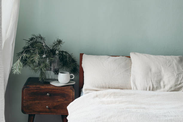 Christmas still life. Cup of coffee, retro wooden bedside table. Pine tree branches and silver stars ornaments in glass vase. Bed with beige cotton muslin beddings, blank sage green wall background.