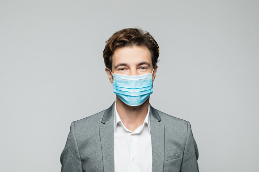 Portrait of pleased young businessman wearing white shirt, grey jacket and N95 face mask looking at camera. Studio shot, grey background.