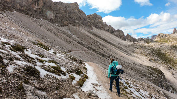 istock A woman with backpack and sticks hiking on a narrow path in Italian Dolomites. There are sharp and steep mountains around her. Lots of lose stones. Raw and desolated landscape. Following the pathway 1289881810