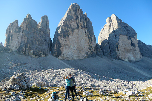 A couple standing on a big boulder and enjoying the close up view on the Tre Cime di Lavaredo (Drei Zinnen), mountains in Italian Dolomites. Sharp and high mountain wall. Desolated and raw landscape