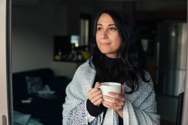 istock Sick Woman holding cup, covered with blanket looking through the window 1289886175