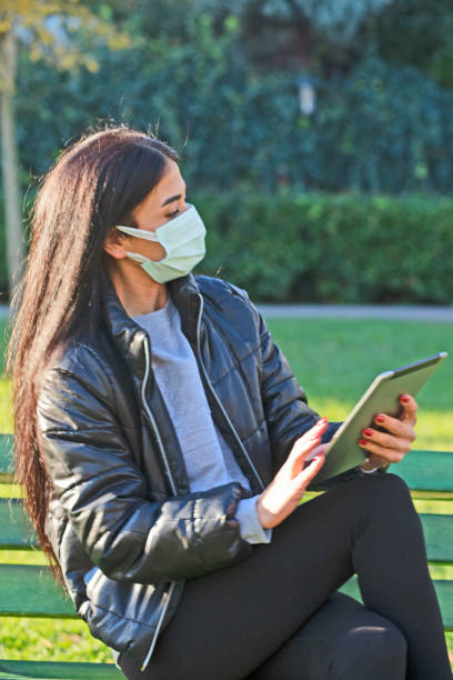 istock Young woman WİTH FACE MASKsitting on a bench in a public park using digital tablet 1289887675