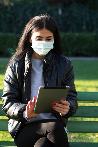 istock Young woman WİTH FACE MASKsitting on a bench in a public park using digital tablet 1289887866