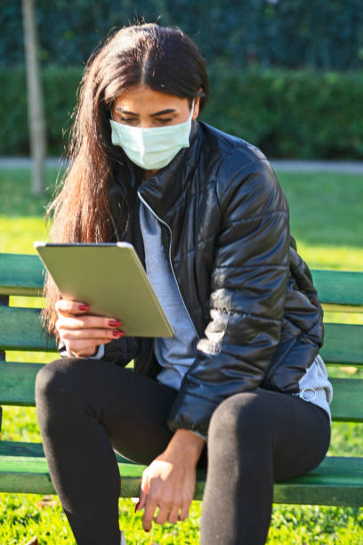 istock Young woman WİTH FACE MASKsitting on a bench in a public park using digital tablet 1289887957