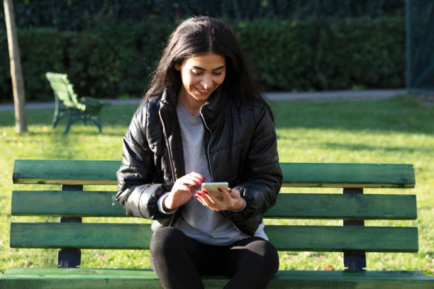 istock Young woman sitting on a bench in a public park using mobile phone 1289888827