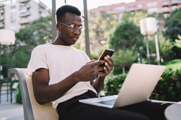 istock Concentrated african american hipster guy in eyewear for vision correction reading notification on smartphone, serious dark skinned businessman checking mails on mobile phone on remote job 1289894291