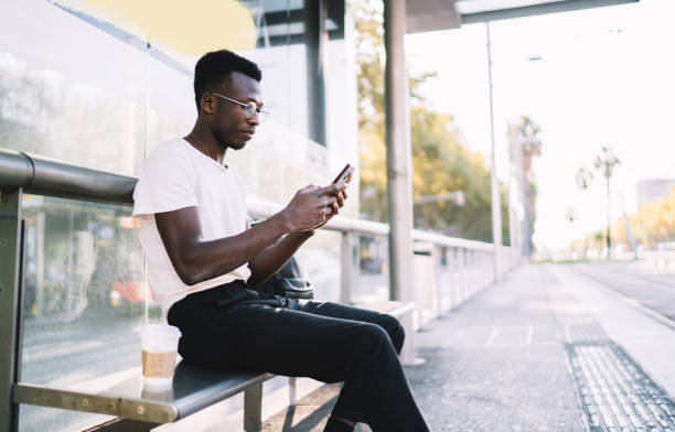 istock Stylish black guy sitting on bus stop with smartphone and coffee 1289894329