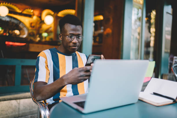 uomo nero intelligente con smartphone e laptop in street café - surfing the net foto e immagini stock