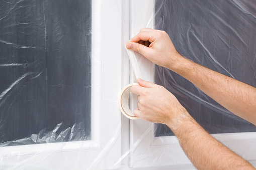 Young adult man hands using paper scotch and gluing plastic film on window frame. Closeup. Preparation before repair work of home.