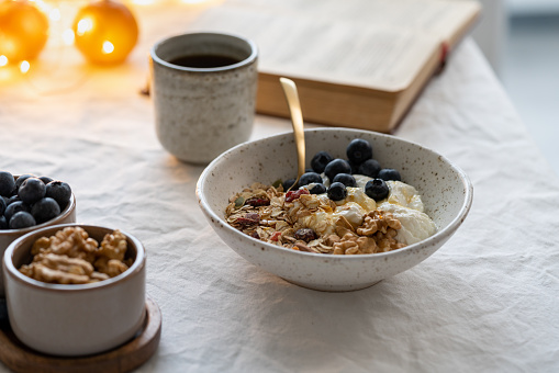 Reading book and eating healthy Christmas holiday winter breakfast with granola muesli and yogurt in bowl on white table background, lights garlands in bokeh. Organic morning diet meal with oat